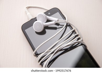 Smartphone With Connected Earphones, White In Color. Earphone Cord Wrapped Around The Phone. Using Earphones To Listen To Music Or To Make Phone Calls.
