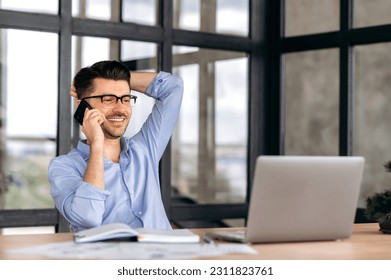 Smartphone conversation. Satisfied friendly caucasian man, office worker or entrepreneur, sitting at a table in the office, having a pleasant phone conversation while working, smiling - Shutterstock ID 2311823761