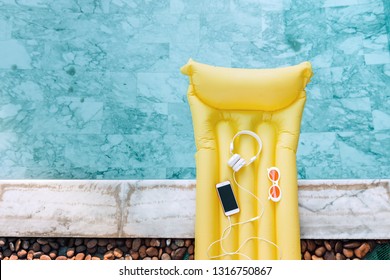 Smartphone, earphones and sunglasses on yellow pool float in blue water, high view from above.