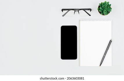 Smartphone with empty display, blank notepad, glasses and green home plant on light background