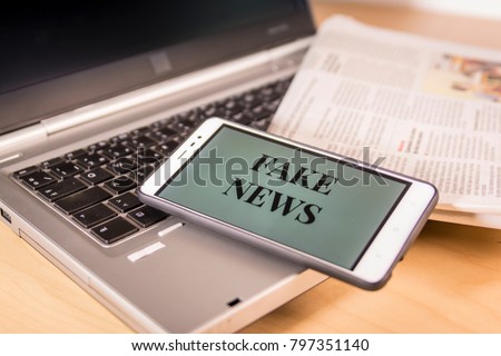 Smartphone with Fake News words on screen over a newspaper and a laptop. HOAX and Fake news concept.