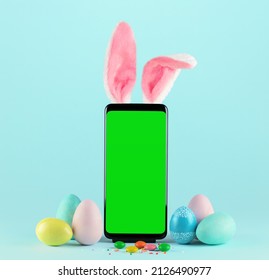 Smartphone with green screen and Easter Bunny ears surrounded by painted eggs on blue background. - Shutterstock ID 2126490977