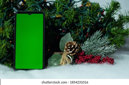 Smartphone With Green Screen On Mobile Phone Near Branch Of Christmas Tree On Background Of A Christmas Tree With Holiday Lights, Viewing A Chroma Key. Online Shopping, Sales