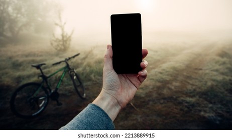 Smartphone Isolated In Hand Against The Background Foggy Morning Landscape And Bicycle. Man Hand Holding Smartphone. Morning Bike Rides.