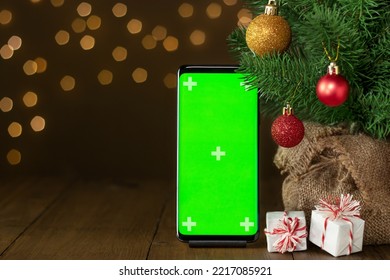 Smartphone Mock Up With Empty Green Screen At Decorated Christmas Tree On Wooden Table With Gift Boxes. Layout For Advertising Products And Services In Mobile Application. Golden Lights In Background.