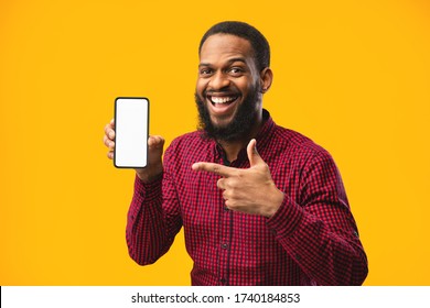 Smartphone Mock Up. Excited casual black man pointing at blank white cell phone screen, empty space, yellow studio