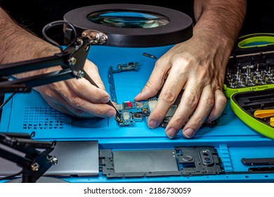 Smartphone Repair In A Service Center. Hands Of A Professional Master Repairman Repairing Disassembled Into Pieces Cell Phone. Internal Parts, Chips, Motherboards, Wiring, Fixing Process.