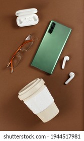 Smartphone, reusable coffee cup, wireless earphones and glasses on brown background top view
