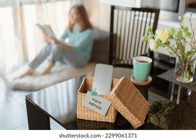 Smartphone is in separate wicker box with inscription digital detox on table. Woman reading book in background. Stop using digital gadgets. Mental and digital detox concept
