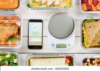 Smartphone With A Weight Loss App And A Food Scale On The Wooden Surface. Lean Fish, A Vegetable Roll, Cashews And Hazelnuts, Salad, A Pie, Chicken And Fresh Fruit. Count Calories.