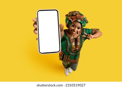 Smartphone with white blank screen in pretty black woman hand, happy young lady wearing national african costume showing great online deal and peace gesture, yellow studio background, mockup, top view - Shutterstock ID 2310519027