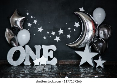 Smashed first birthday white cake with stars and one candle for little baby boy and decorations. Black background. Big silver letters ONE, silver stars and different balloons.