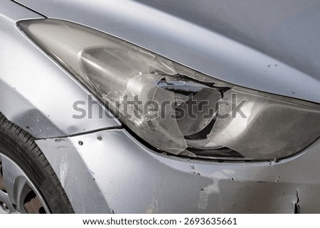 smashed headlight and surrounding damaged bumper of a silver car, with cracks clearly fracturing the lens. The visible scuff marks a localized frontal impact requiring immediate repair.