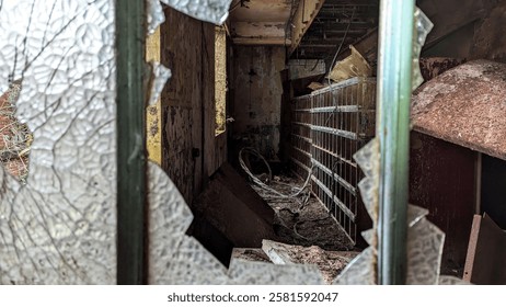 Smashed window, broken glass looking through into derelict spooky room of abandoned building. Rusted shattered frame with peeling paint and mould. Dark creepy dusty and cluttered storeroom with natural light and shadow from missing pane. Aftermath of vandalism and wreckage of disrepair.