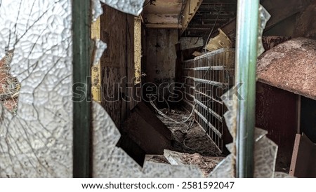 Smashed window, broken glass looking through into derelict spooky room of abandoned building. Rusted shattered frame with peeling paint and mould. Dark creepy dusty and cluttered storeroom with natural light and shadow from missing pane. Aftermath of vandalism and wreckage of disrepair.