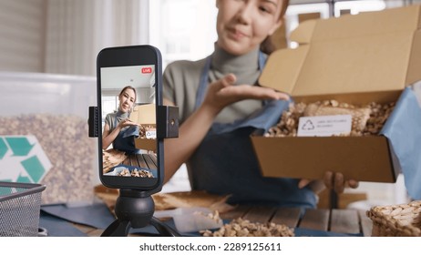 SME owner go green net zero retail store live stream talk on phone showing eco care plastic free packaging box. Asia people young woman record video on tiktok IG reel instagram shop happy side hustle.