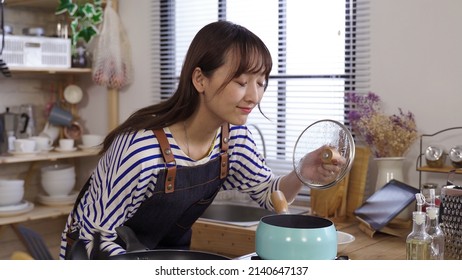 Smelling The Nice Aromas From Her Meal. Portrait Happy Japanese Lady Bending Toward Stove And Lifting The Lid Off Kitchen Pot. Real Moments