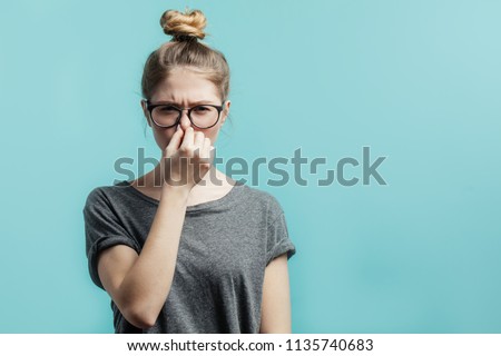 Smells bad, awful odor. Young student girl with hair bun in casual t-shirt squeezing nose with fingers, looking in camera with disgust expression, eager to slip away from bad smell indoors