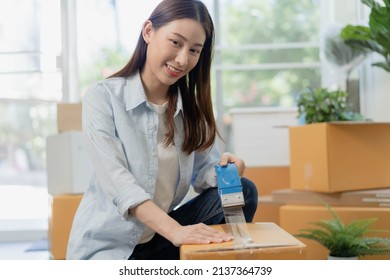 Smile Asian Young Woman Packing, Box By Tape Machine, Sealing Cardboard Indoors. Business Owner In Warehouse, Moving Or Preparing Move In New House, Apartment. Relocation Concept.