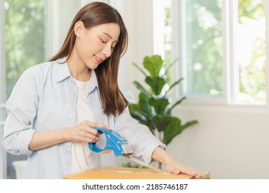 Smile Asian Young Woman Packing, Box By Tape Machine, Sealing Cardboard Indoors. Business Owner In Warehouse, Moving Or Preparing Move In New House, Apartment. Relocation Concept.