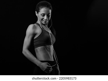 Smile athlete female model in sport bra listening music in headphones standing and looking down on black background with empty space. Healthy lifestyle portrait. 
