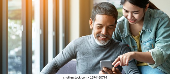 Smile attractive stylish short beard mature asian man using smartphone with young woman. Daughter teach asian old man or dad using internet social media network technology with digital gadget at home.