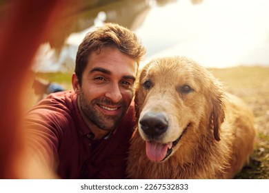 Smile, boy. Cropped shot of a handsome young man and his dog taking selfies by a lake in the park. - Powered by Shutterstock