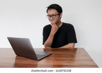 Smile Face And Funny Tricky Awkward Smirk Face Of Young Asian Man With Hand In Chin In A Black T-shirt In A Front Of A Laptop.