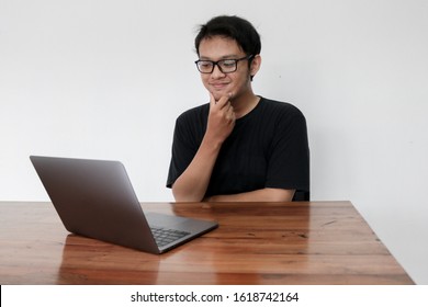 Smile Face And Funny Tricky Awkward Smirk Face Of Young Asian Man With Hand In Chin In A Black T-shirt In A Front Of A Laptop.