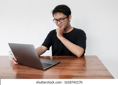 Smile Face And Funny Tricky Awkward Smirk Face Of Young Asian Man With Hand In Chin In A Black T-shirt In A Front Of A Laptop.