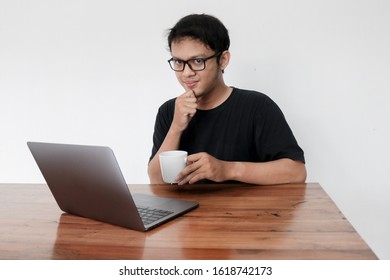 Smile Face And Funny Tricky Awkward Smirk Face Of Young Asian Man With Hand In Chin In A Black T-shirt In A Front Of A Laptop.