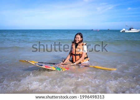 smile happy Asian woman with life-saving jacket sit on small paddleboard holding oar at wavy sea beach, girl relax on vacation in summer