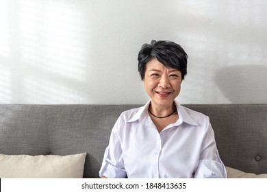 Smile and happy Middle-aged gray-haired Asian woman wearing white shirt pose at home on a sunny morning, a single retired woman sitting on the sofa in the living room. Lonely Elder Concept. - Powered by Shutterstock