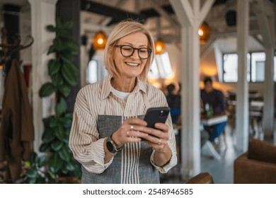 smile mature woman in apron stand at restaurant and use mobile phone, beautiful woman owner stand and use mobile phone at restaurant