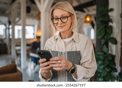smile mature woman in apron stand at restaurant and use mobile phone, beautiful woman owner stand and use mobile phone at restaurant