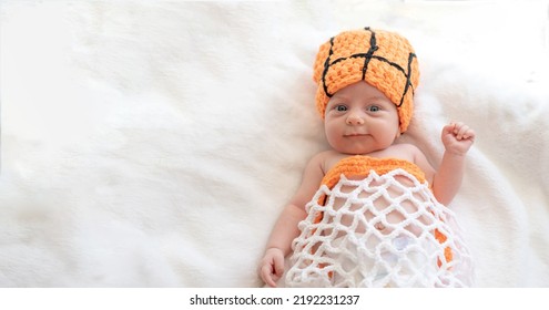 Smile Newborn Baby With Blue Eyes Lying On His Back On White Sheet With Orange Hat As A Ball, And Clothes As A Basket Net, Playing Basketball. Top View. Space For Text