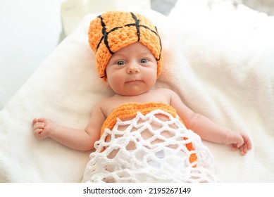 Smile Newborn Baby With Blue Eyes Lying On His Back On White Sheet With Orange Hat As A Ball, And Clothes As A Basket Net, Playing Basketball. Top View. Companion Group Game