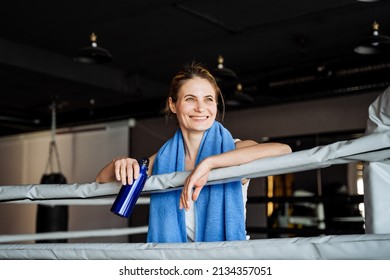 A smile on the young girl's face. Blue water bottle in hand. Rest after a power load in the gym. Fitness model in the boxing ring. Towel around the neck. High quality photo - Powered by Shutterstock