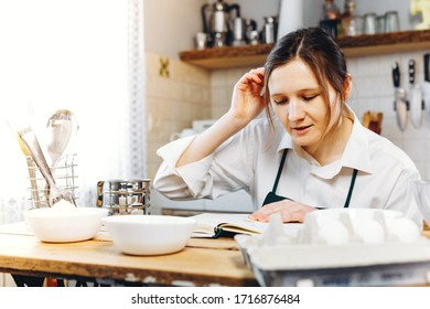 Smile Woman Sitting At Wooden Kitchen Table With Recipe Book And Trying To Choose What To Cook. Cooking At Home Concept, Lifestyle. Ketogenic Diet And Menu