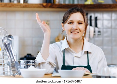 Smile Woman Sitting At Wooden Kitchen Table With Recipe Book And Trying To Choose What To Cook. Cooking At Home Concept, Lifestyle. Ketogenic Diet And Menu