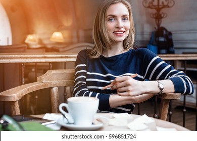 A smile young woman dreams seat in a cafe with cup of coffee with wooden pencil in hand. Concept of planning personal training schedule.Looking at camera.