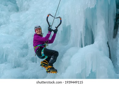 Smiled Caucasian Woman Hiking At A Frozen Waterfall, Holding The Ice Axes In Heart Shape And Hanging On A Climbing Rope 