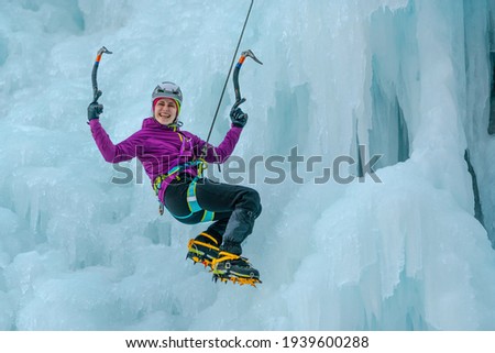 Smiled Caucasian woman hiking at a frozen waterfall, holding the ice axes and hanging on a climbing rope 
