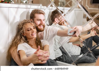 Smiled couple making a selfie sitting on the floor near the table. Blonde man and woman in white T-shirts.