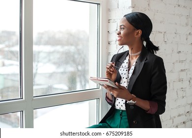 Smiled Girl With Dark Shiny Skin Sitting On The Windowsill, Holding The Tablet And Looking Out The Window.