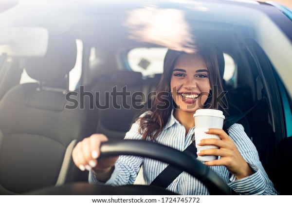 Smiled woman sitting in the car holding a cup of\
coffee. She has a beautiful toothy smile. Success businesswoman\
traveling by car with coffee to go. Woman driving and holding a\
coffee cup