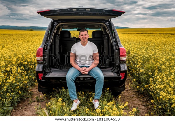 smiled young man sitting in the trunk of a car in\
yellow rape field