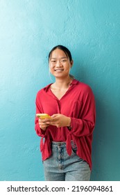 Smiley Asian Office Worker Holding Her Mobile Phone And Looking At Camera Against Blue Background.