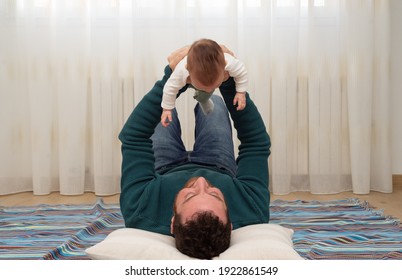 Smiley Father Lying On A Blue Blanket While Is Lifting His Baby Into Air. 