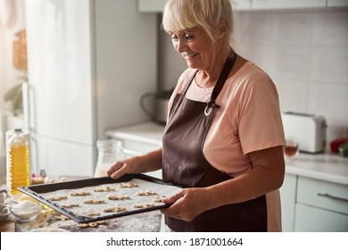 Smiley grey-haired woman wearing apron and holding a baking sheet full of dough shapes